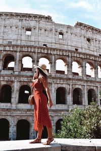 Full length of woman in dress standing on retaining wall against coliseum during sunny day