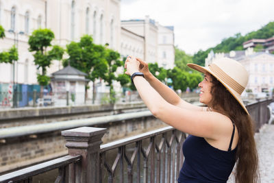 Side view of young woman holding hat standing against built structure