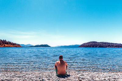 Rear view of shirtless mid adult man sitting at beach against clear blue sky