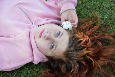 A red-haired girl in a pink hoodie lying on the grass looks at you with blue eyes