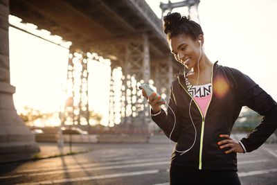 Happy female jogger using phone while standing on city street