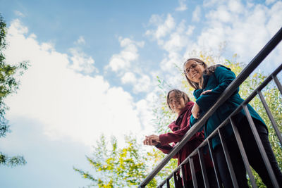 Low angle view portrait of sisters standing by railing on bridge against sky