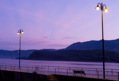 Street lights by lake against sky during sunset