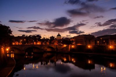 Illuminated buildings by river against cloudy sky during night