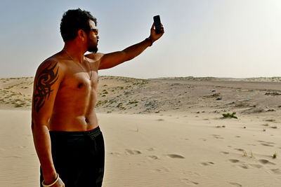 Shirtless young man taking selfie with mobile phone at desert during sunny day