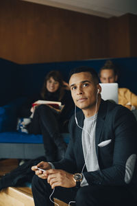 Mid adult businessman looking away while listening to in-ear headphones sitting against colleagues at office cafeteria