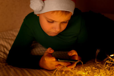 A child in a red cap lies on the bed and swipes in a smartphone. a boy on christmas night looks