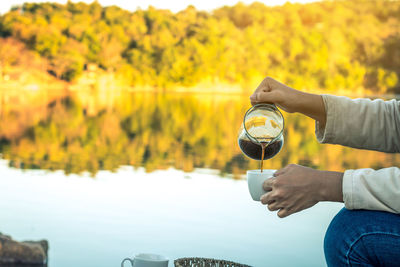 Close-up of hands pouring drink in cup against trees against sky