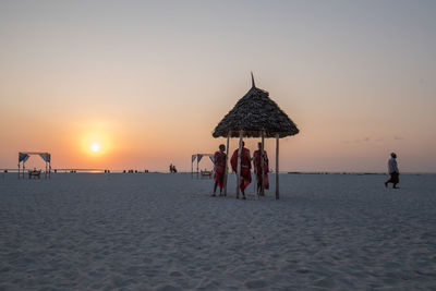 People standing in thatched roof at beach during sunset