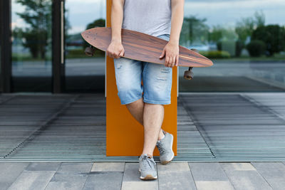 Close up outdoor portrait of young man in shorts and grey sneakers holding longboard in hands