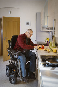 Full length of retired senior man with disability sitting in motorized wheelchair cutting bell pepper at kitchen counter