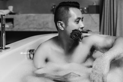Shirtless man carrying rose in mouth while siting in bathtub