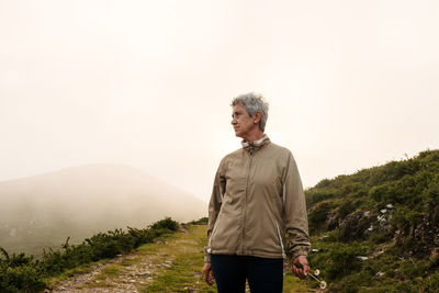 Aged female traveler with short gray hair looking away and walking on path near hill at daytime in nature