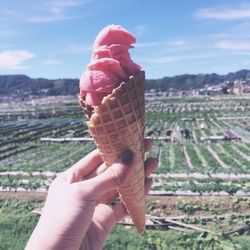 Cropped hand of woman holding ice cream cone over field against sky