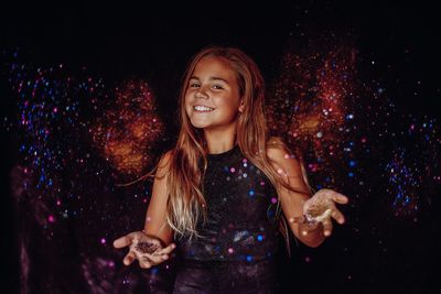 Portrait of smiling girl throwing glitter against black background