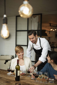 Smiling man wearing apron serving wine to businesswomen at table