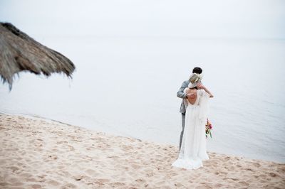 Romantic newlywed couple standing at beach against sky
