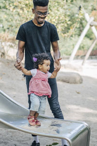 Young man holding hands of daughter standing on slide at playground