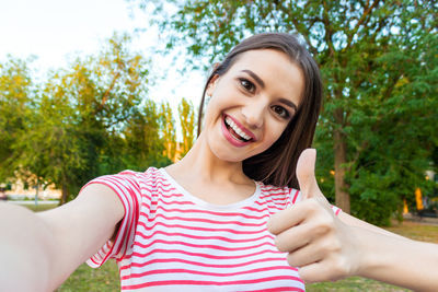 Portrait of smiling beautiful woman showing thumbs up sign at park