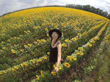 Young woman in a black dress and hat stands on  field with sunflowers in summer and takes a selfie