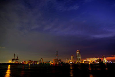 Illuminated factory by sea against sky at night