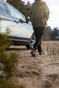 Young no face girl in green jacket, black jeans and brown boots walking on beach along car.