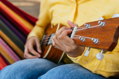 Cropped unrecognizable female in bright shirt lounging in park hammock and playing ukulele