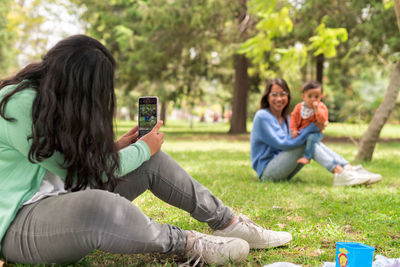 Young female taking photo on cellphone of cheerful mom raising baby while sitting on lawn in park