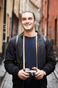 Portrait of smiling young man holding camera while standing in alley