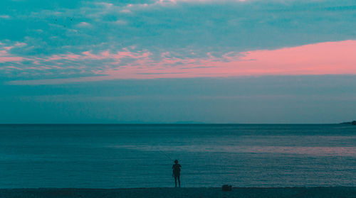 Silhouette woman fishing on shore at beach against sky during sunset