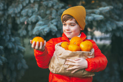 A boy in a bright orange jacket and yellow hat holds a large bag of oranges in his hands