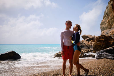 Young man and young woman dressed in swimsuits hug standing on a beach opposite surfing waves