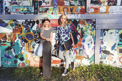 Full length of girls holding skateboards while standing against graffiti wall