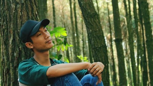 Thoughtful young man by tree trunk sitting in forest