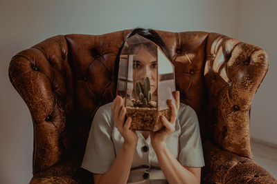 Tranquil female sitting in shabby leather armchair and looking at camera though glass pot with green cactus