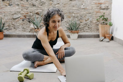 Smiling woman using laptop for learning yoga through online tutorial while sitting at back yard