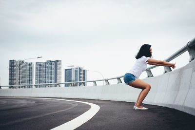 Full length side view of woman exercising by railing on bridge in city against sky