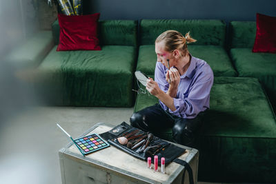 Man applying make-up looking in hand mirror sitting on sofa