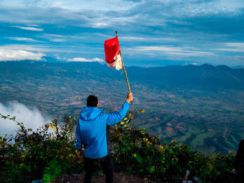 Rear view of man holding flag on cliff against mountain and sky