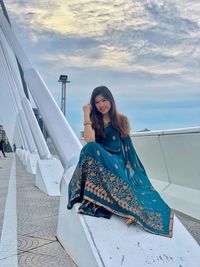 Full length portrait of smiling woman sitting on cable-stayed bridge against sky