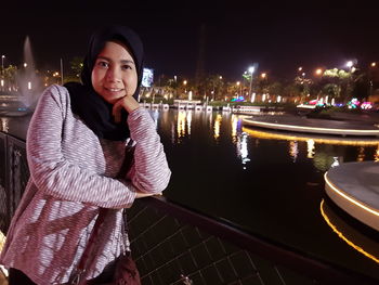 Portrait of smiling young woman standing by river in city at night