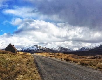 Country road leading towards mountains against cloudy sky