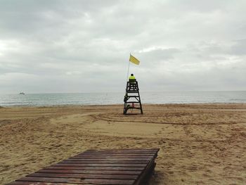 Rear view of man sitting on lifeguard chair at beach against cloudy sky