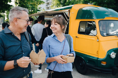 Happy male and female friends talking while standing with box against commercial land vehicle