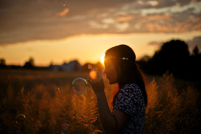 Side view of woman blowing bubbles while standing on field during sunset