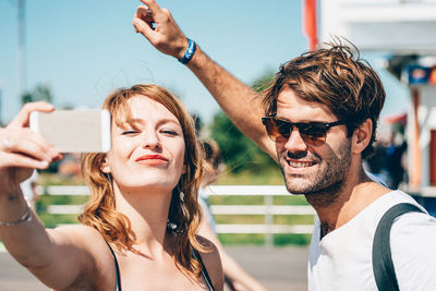 Happy friends clicking selfie on smart phone while standing outdoors