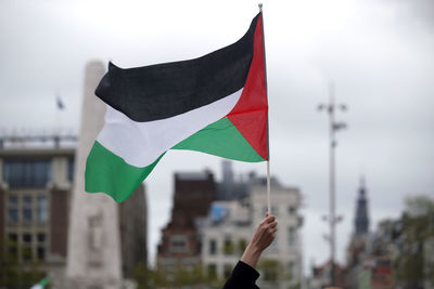 Cropped image of hand holding flag against sky in city