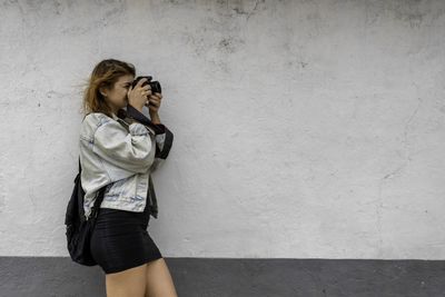 Side view of woman photographing while standing against wall outdoors