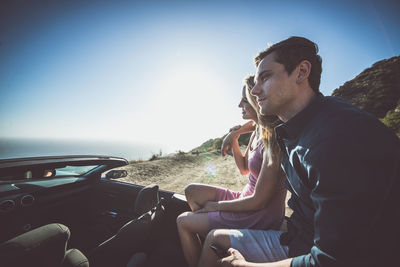 Young man sitting on car against sky