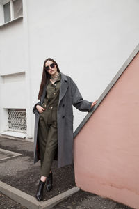 Full length front view portrait of young woman in grey coat and suit against wall outdoors, outwear 